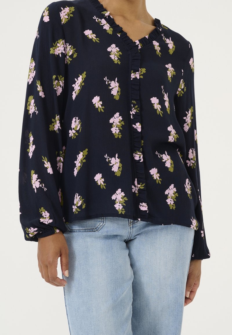 Femme portant un chemisier bleu marine à imprimé floral rose avec un devant à volants, associée à un jean en denim bleu clair, debout devant un fond uni.