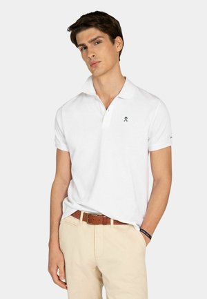 Joven vestido con una camisa polo blanca de manga corta con un pequeño logo, pantalones beige, cinturón marrón y pulsera negra, posando frente a un fondo liso.