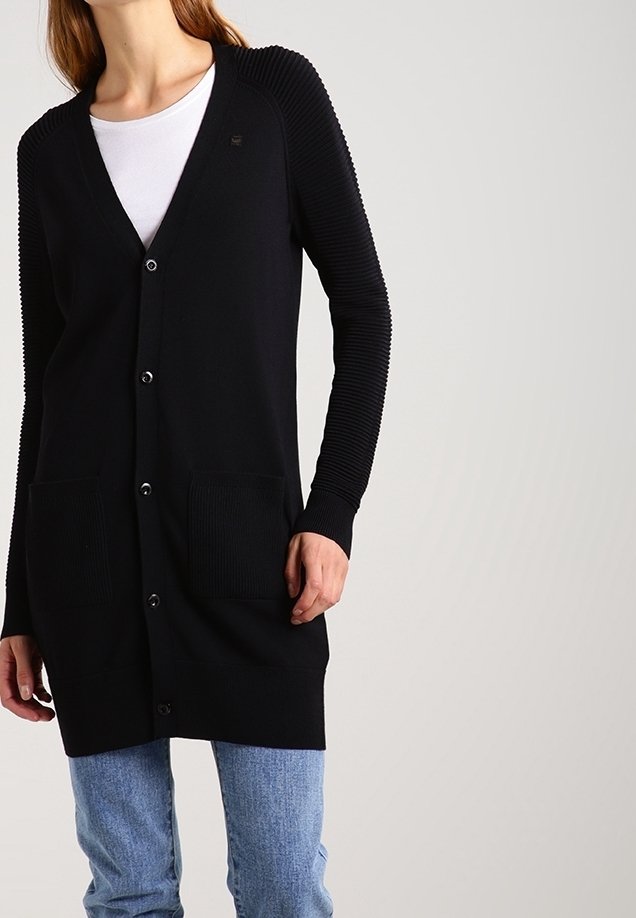 Femme portant un long cardigan noir boutonné avec des manches côtelées et des poches, par-dessus une chemise blanche et un jean bleu, sur un fond uni.