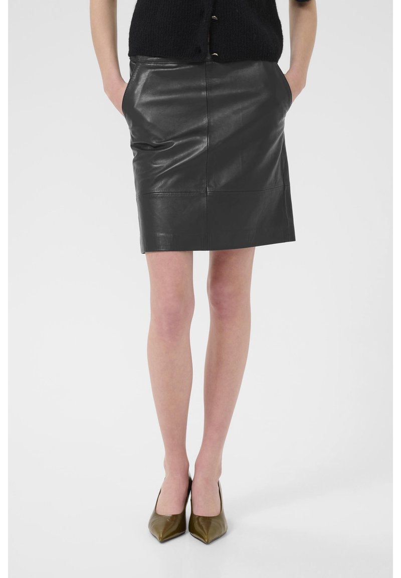 Femme portant une jupe en cuir noire avec des poches, un haut noir et des talons pointus vert olive, debout les mains dans les poches.