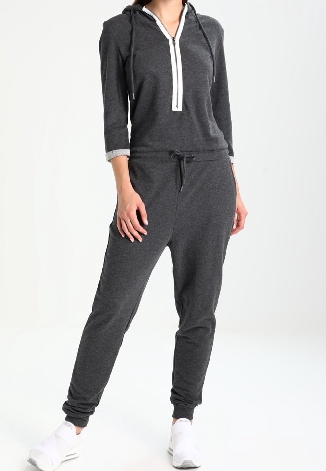 Personne portant une combinaison à capuche gris foncé avec fermeture éclair à l'avant et des baskets blanches, debout avec une jambe légèrement en avant.