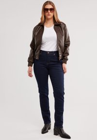 Noor naine, kes kannab tumepruuni nahktagi, valget särk, tumedat sinist teksaseid, musti kontsakingi ja suurte prillidega, seisab heletaustal.