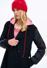 Žena s červenou pletenou čepicí, černým kabátem s vycpávanými rukávy, růžovou mikinou a vícebarevným náhrdelníkem z korálků, která se dívá stranou.