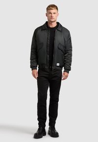 Jachetă neagră tip bomber cu o finisare netedă, închidere cu fermoar și două buzunare frontale. Pantaloni negri slim-fit și cizme negre cu șireturi completează ținuta.