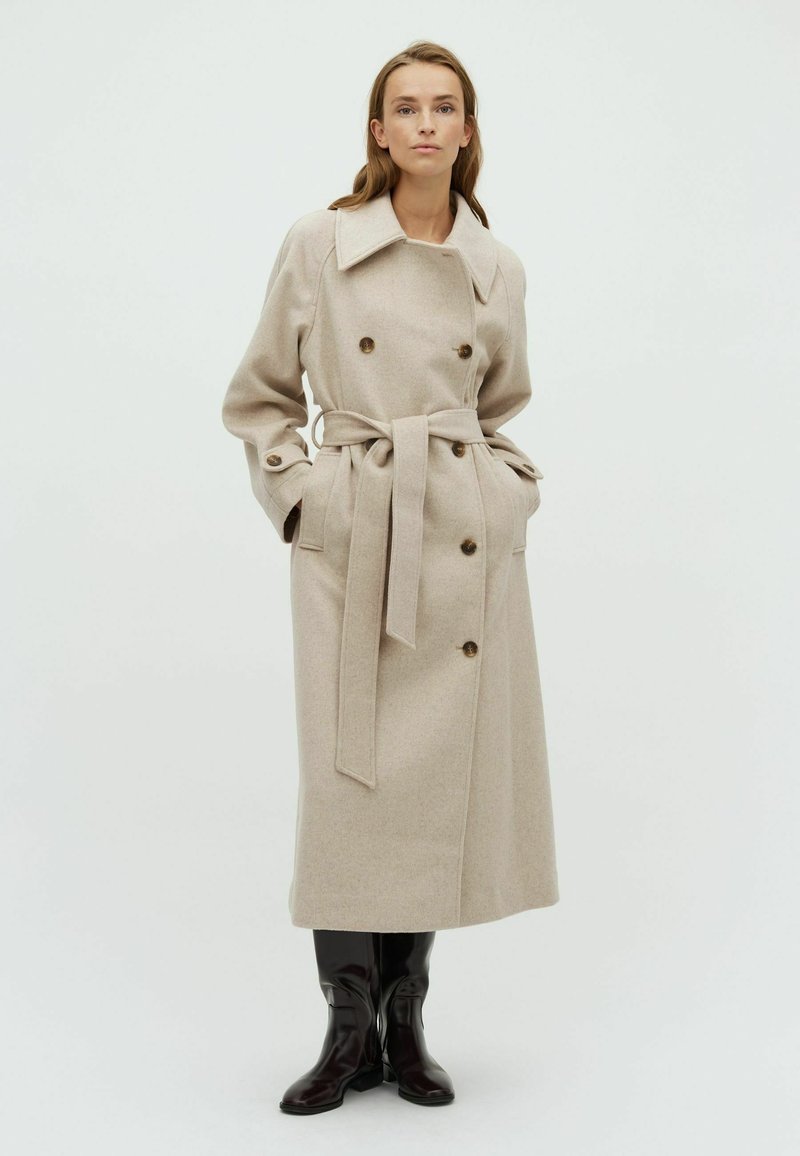 Cappotto trench in lana beige con design doppiopetto, grande collo, vita con cintura e tasche laterali. Bottoni marroni lucidi aggiungono un tocco di dettaglio.