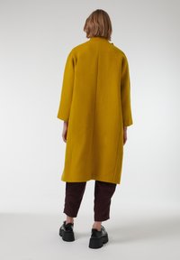 Abrigo de lana amarillo con mangas largas, silueta recta y bolsillos laterales, combinado con pantalones oscuros y zapatos de plataforma.