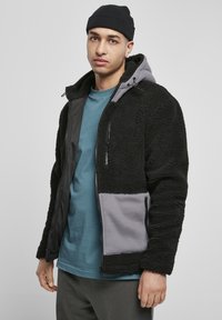Jachetă din fleece neagră cu accente gri, având un fermoar în față, buzunare laterale și glugă. Purtați deasupra unei cămăși albastre și a unor pantaloni gri.