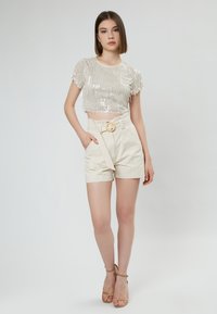 Top a maniche corte in paillettes argento, accostato a pantaloni corti beige a vita alta con cintura e fibbia circolare.