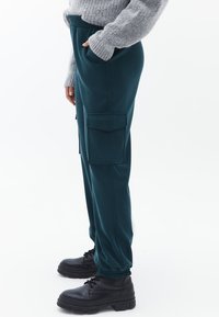 Pantaloni cargo verde petrolio con vita elastica e polsini alla caviglia, dotati di due tasche laterali e di una texture liscia, abbinati a stivali neri con lacci.