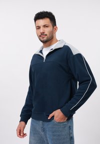Uomo dai capelli scuri e ricci che indossa un pile blu navy e grigio con zip e jeans blu, in piedi con una mano in tasca contro uno sfondo bianco.