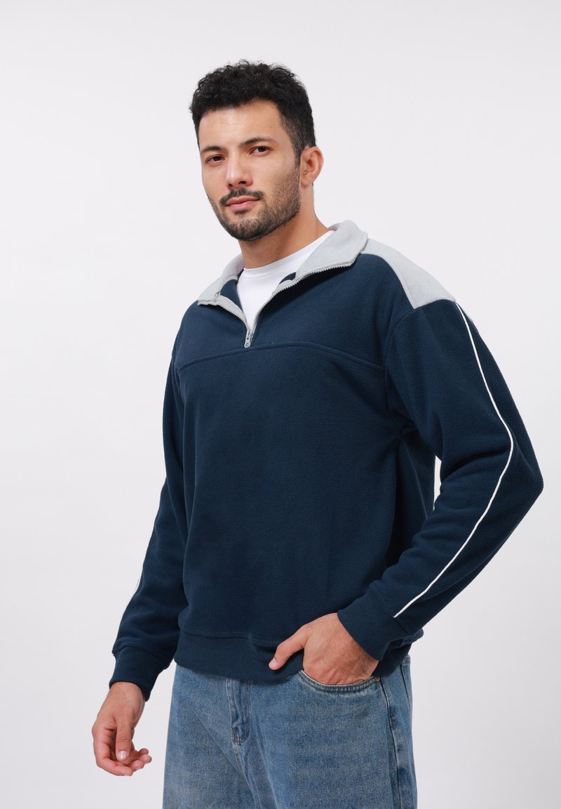 Uomo dai capelli scuri e ricci che indossa un pile blu navy e grigio con zip e jeans blu, in piedi con una mano in tasca contro uno sfondo bianco.