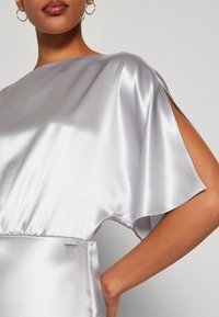 Robe en satin argenté avec un large décolleté et des manches évasées. Présente une taille froncée et une petite étiquette métallisée de marque sur le côté.