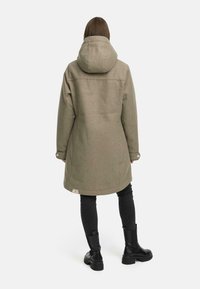 Olivengrønn ullfrakk med hette, med rett snitt, knappedede ermer og en logomerking nær kanten. Båret over svarte bukser.