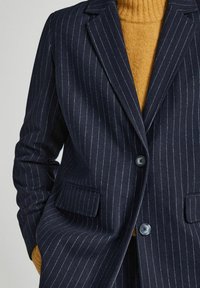 Blazer de rayas marineras con dos botones, solapas normales y bolsillos laterales. Combinado con un suéter de cuello alto de canalé en color mostaza.