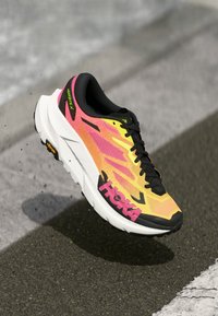 Sapatilha de corrida laranja brilhante e amarela com o logótipo rosa "HOKA" no ar sobre a superfície de uma estrada de asfalto.