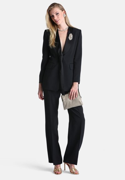 Femme en costume noir avec un décolleté plongeant, tenant un sac à main argenté, portant des sandales à talons hauts dorées et des bijoux argentés, incluant des boucles d'oreilles et un collier.