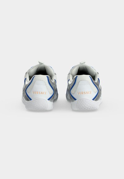 Paire de baskets blanches et argentées avec des accents bleus, montrant la vue arrière avec le logo "VERSACE" en or sur chaque talon.