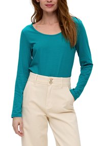 Tefarbenes Langarmshirt mit Rundhalsausschnitt, kombiniert mit hochtaillierten cremefarbenen Hosen mit zwei Knöpfen und seitlichen Taschen. Glatte Stofftextur.