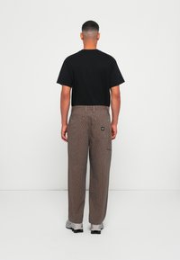 Pantaloni a righe marroni con una vestibilità rilassata, dotati di tasche posteriori, indossati con una maglietta nera e sneakers grigie.
