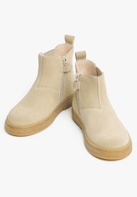 Stivaletti alla caviglia color beige in suede, con suola piatta e texture, cerniera laterale e linguetta per il sollevamento. Dettagli di cucitura visibili lungo le cuciture per un design aggiuntivo.