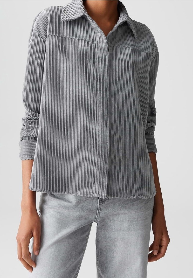 Personne portant une chemise en velours côtelé gris clair à manches longues avec des rayures verticales et un jean gris clair, debout devant un fond uni.