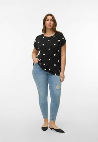 Vrouw met een zwart T-shirt met korte mouwen en crèmekleurige hartjes, lichte ripped skinny jeans en zwarte, puntige instapschoenen, staand op een witte achtergrond.