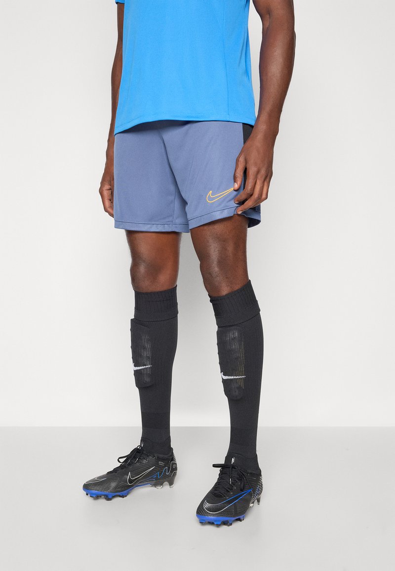 Conjunto deportivo de Nike que incluye una camiseta azul, pantalones cortos grises con logo amarillo, calcetas negras y zapatillas negras con acentos azules.