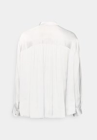 Blusa bianca a maniche lunghe con schiena plissettata, colletto e polsini con bottone. Tessuto liscio con un leggero lucido, caratterizzato da una vestibilità sartoriale.