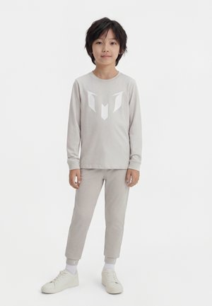 Barn med mørkt hår iført lysegrå langærmet skjorte og bukser med hvidt logo, hvide sokker og hvide sneakers, stående på hvid baggrund.