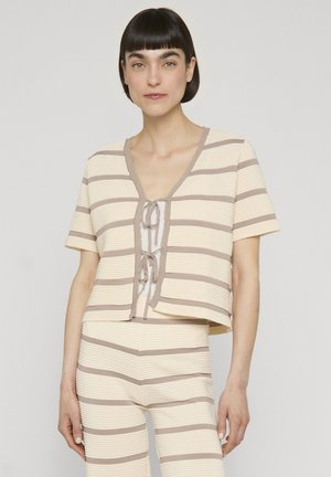 Kvinde med kort mørkt hår iført beige og taupe vandrette stribede kortærmede top med knuder foran og matchende bukser mod ensfarvet baggrund.