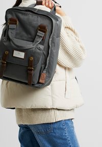 Sac à dos en toile grise avec des accents en cuir brun, de forme rectangulaire, poche avant et deux poignées supérieures. Porté par-dessus un gilet matelassé de couleur claire.