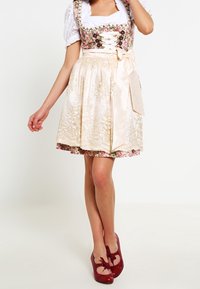 Dirndl con motivo floreale, corpetto allacciato, gonna in raso color crema e nastro annodato in vita. Abbinato a scarpe rosse con fiocchi.