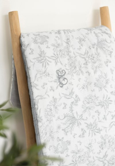 Πάπλωμα με ραμμένα μοτίβα λουλουδιών σε γκρι χρώμα πάνω σε λευκό φόντο, με γκρι υφασμάτινη περίμετρο, κρεμασμένο σε ξύλινη σκάλα.