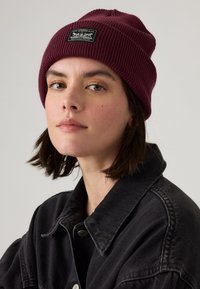 Burgunderribbet strikket hue med en sort label med hvid tekst; bæres med en sort denimjakke. Glat tekstur og tætsiddende pasform.