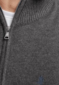Gros plan sur un pull-over gris en tricot zippé avec une chemise blanche en dessous et une petite figure brodée sur la poitrine.