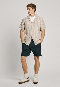 Camisa de manga corta beige con rayas verticales; combinada con pantalones cortos color marino. Zapatos blancos con suelas marrones, usados con calcetines de color claro.