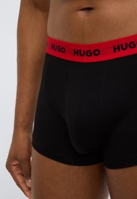 Μαύρα ανδρικά εσώρουχα boxer με κόκκινη μέση που φέρει την επωνυμία "HUGO" με μαύρα γράμματα. Κατασκευασμένα από λείο, ελαστικό ύφασμα.