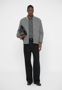 Γκρίζα μάλλινη bomber ζακέτα με μαύρες και άσπρες ρίγες στους καρπούς, φορεμένη πάνω από έναν υφασμάτινο γκρίζο πουλόβερ, σε συνδυασμό με μαύρες φαρδιές παντελόνες και δερμάτινα παπούτσια.