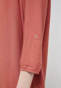 Rustfarbenes Hemd aus strukturiertem Stoff, mit Dreiviertelärmeln und einem Knopfdetail an den Manschetten. Einfaches und klares Design.