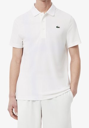Uomo che indossa una polo bianca a maniche corte con un piccolo logo verde a forma di coccodrillo sul petto sinistro e pantaloni bianchi abbinati.