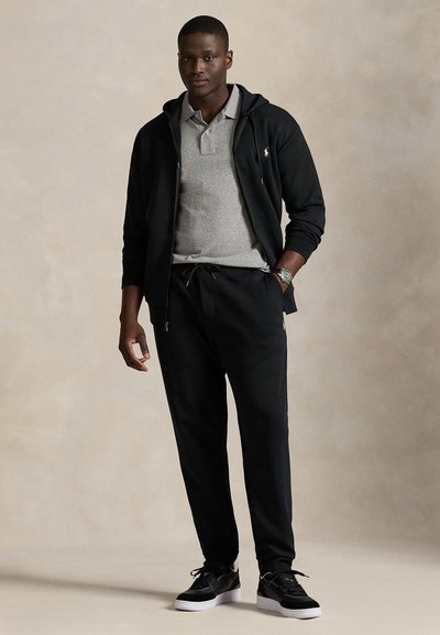 Svart huvtröja med grå pikétröja under, kombinerad med svarta mjukisbyxor och svarta sneakers. Tyget verkar mjukt med en avslappnad passform.