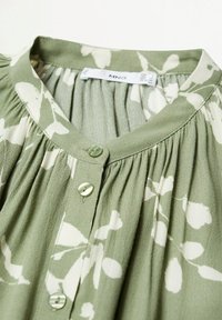 Blusa verde con patrón floral blanco, con escote abotonado, textura de tela plisada y material suave y ligero.