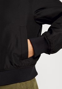 Main d'une personne partiellement dans la poche latérale d'une veste noire avec des poignets et une taille côtelés, associée à un pantalon vert olive.