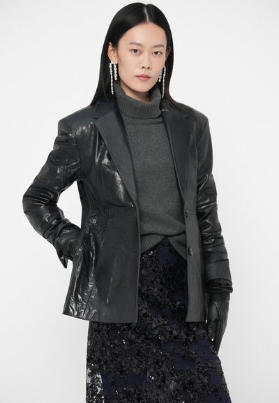 Blazer de couro preto com um acabamento brilhante, combinado com um suéter de gola alta cinza-escuro e uma saia preta de lantejoulas, destacando a textura e as sobreposições.