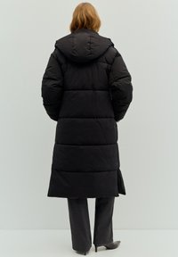 Schwarzer Steppmantel mit Kapuze, lang geschnitten, mit gestepptem Design und elastischen Bündchen, getragen über grauen Hosen und Schuhen mit Absatz.