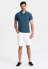 Námořnická modrá polo košile s krátkým rukávem a jemným logem. Bílé kraťasy a černé nazouvací boty. Čistý, minimalistický design a střih.