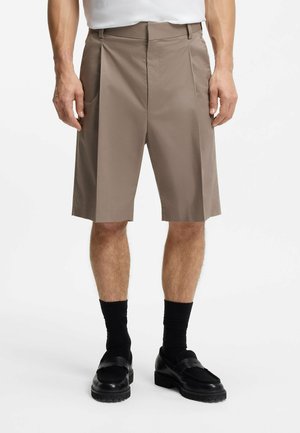 Mann trägt taupefarbene, knielange, maßgeschneiderte Shorts, schwarze Socken und schwarze Loafers, steht vor einem schlichten weißen Hintergrund.