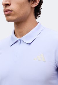 Jeune homme portant un polo zippé violet clair avec un logo triangulaire sur la poitrine gauche, vu du cou au haut du torse.
