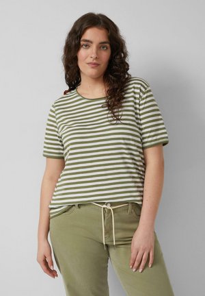 Kobieta z długimi kręconymi włosami, ubrana w zielono-białą, paskowaną koszulkę i oliwkowo-zielone spodnie ze białym sznurkiem, stojąca na szarym tle.