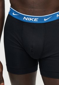 Άνδρας που φοράει μαύρα boxer με μπλε ζώνη μέσης με το λογότυπο της Nike και το σύμβολο swoosh σε απλό φωτεινό φόντο.
